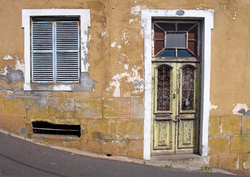 Front of an old abandoned yellow house with blue shuttered windows and locked green wooden door with flaking peeling paint on. The front of an old abandoned stock photos